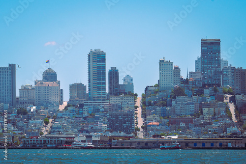 a densely packed San Francisco cityscape with high-rise buildings and residential areas leading up steep hills. The bay is in the foreground, with a clear blue sky above.