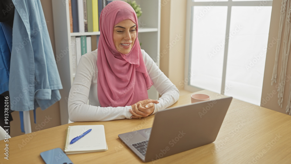 A smiling woman wearing a pink hijab sits at a desk with a laptop in a bright home office setup. A smiling woman wearing a pink hijab sits at a desk with a laptop in a bright home office setup.