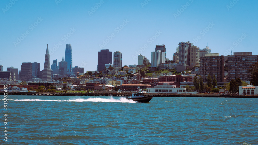 Obraz premium San Francisco skyline, including the Transamerica Pyramid and Salesforce Tower, with a boat speeding across the water in the foreground. The scene is set under a clear blue sky.