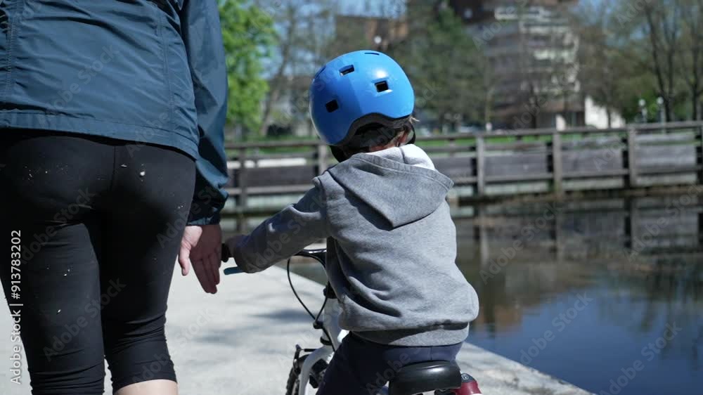 Back of Young boy riding a bike with a blue helmet, learning to balance with an adult's help Back of Young boy riding a bike with a blue helmet, learning to balance with an adult's help