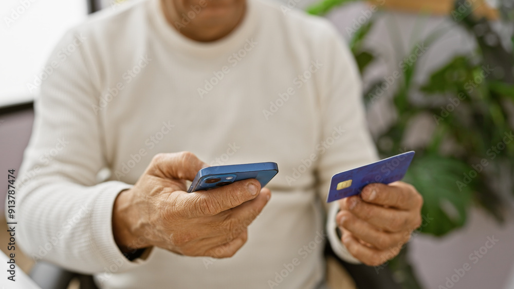 A middle-aged hispanic man using a smartphone and holding a credit card indoors, likely in an office setting. A middle-aged hispanic man using a smartphone and holding a credit card indoors, likely in an office setting.