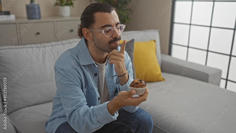 Young man with moustache sitting in living room holding a cupcake and thinking in front of a sofa while wearing a light blue jacket and eyeglasses. Young man with moustache sitting in living room holding a cupcake and thinking in front of a sofa while wearing a light blue jacket and eyeglasses.