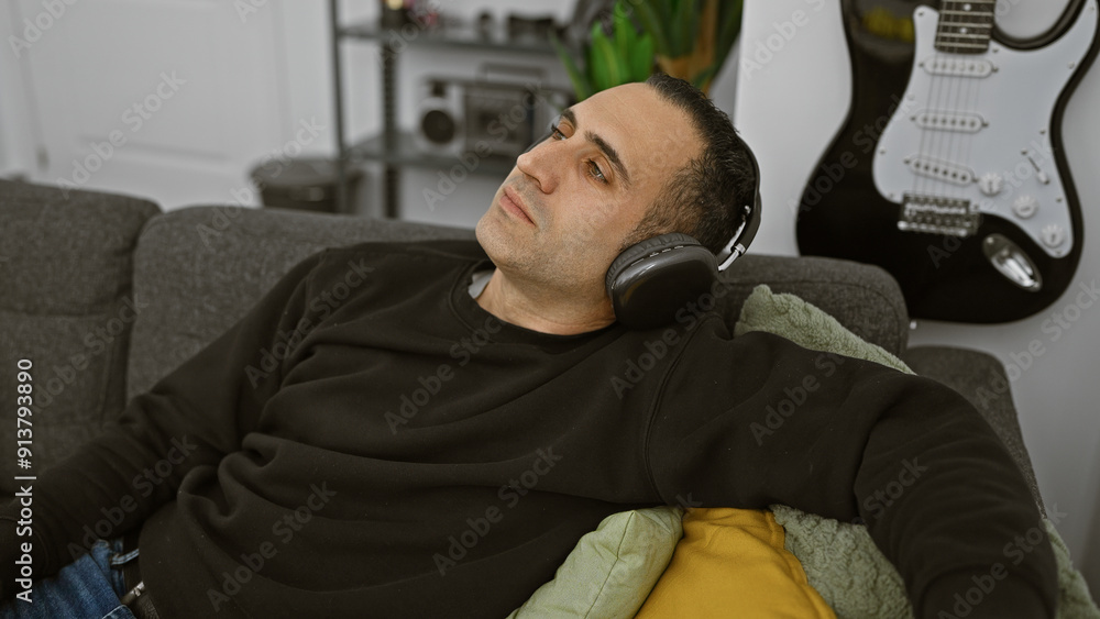 A relaxed hispanic man enjoys music with headphones in a cozy living room, featuring a guitar and a sofa. A relaxed hispanic man enjoys music with headphones in a cozy living room, featuring a guitar and a sofa.