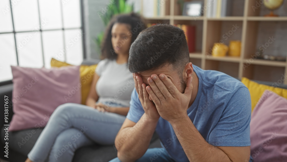 A concerned woman watches as a distraught man covers his face while sitting on a couch in a modern living room. A concerned woman watches as a distraught man covers his face while sitting on a couch in a modern living room.