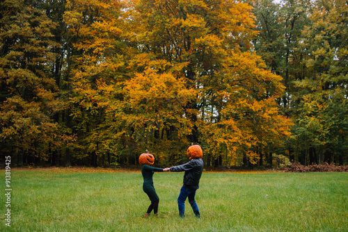 A couple with jack o'-lantern costumes holding each other's hands and dancing on a green lawn against yellow October forest on a background