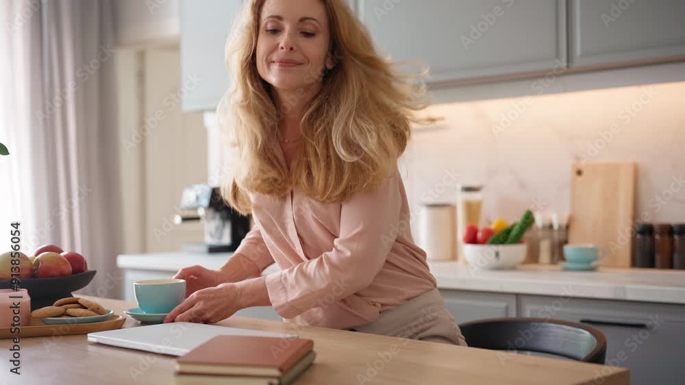 Pretty freelancer carrying cup of tea and siting at table in kitchen. Woman opening top of laptop. Getting ready to start her working day. Freelancing from home. Workplace. Online job.