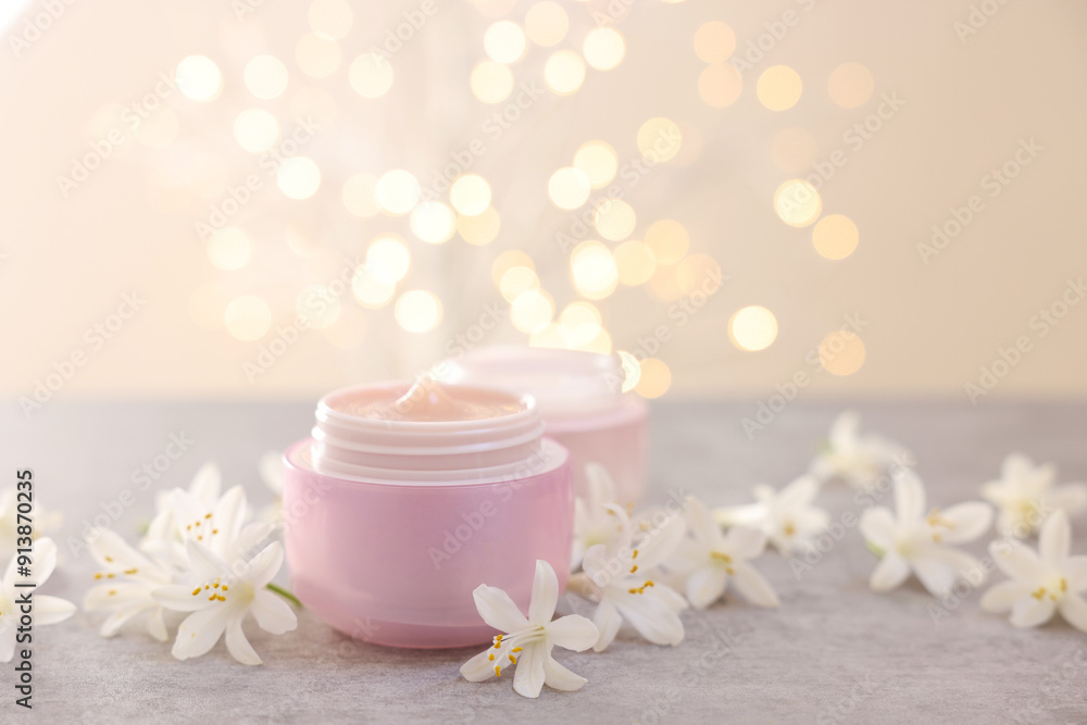 Cream in jars and beautiful jasmine flowers on grey table against beige background with blurred lights, closeup Cream in jars and beautiful jasmine flowers on grey table against beige background with blurred lights, closeup