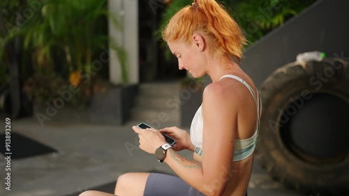 Redhead female athlete checks fitness tracker on wrist in open-air tropical gym. Woman syncs health data to smartphone app post workout, reviews progress in lush palms. Slow motion. photo