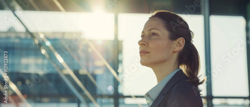 Free Download Professional woman in a business suit standing confidently in a sunlit office, exuding ambition and focus. - GFXLoad