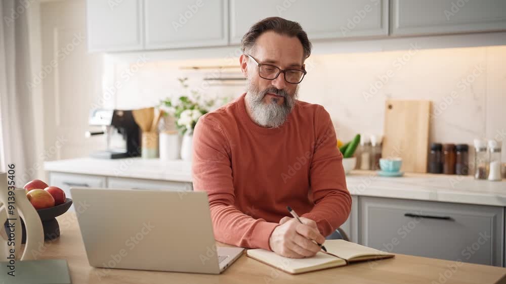 Older Caucasian male with big grey beard enjoying his free time in kitchen. Watching video or reading through recipe. Making notes in his notebook with pen. Using glasses for better vision. Older Caucasian male with big grey beard enjoying his free time in kitchen. Watching video or reading through recipe. Making notes in his notebook with pen. Using glasses for better vision.
