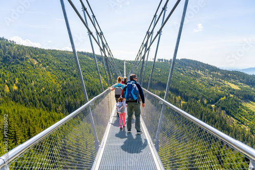 People walk across the long suspension footbridge in Dolni Morava, Czechia. A view of the green forest surrounds the bridge. photo