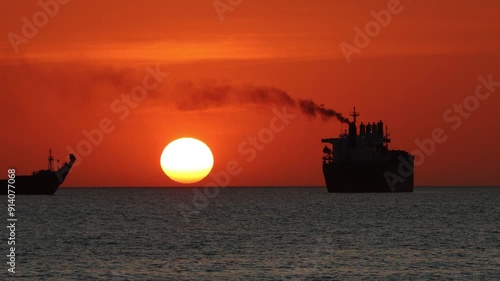 A ship is sailing next to another ship which is anchored in the sea with a sunset in the background