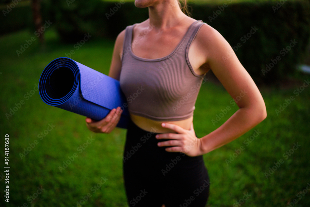 Slim young woman holding rolled fitness mat in park at sunset, wellbeing and healthy sportive lifestyle Slim young woman holding rolled fitness mat in park at sunset, wellbeing and healthy sportive lifestyle