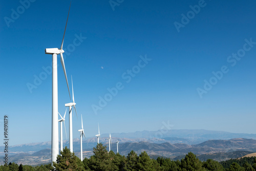 Panoramic View of Wind Turbines on a Mountain Ridge Under a Clear Blue Sky with the Moon Visible in the Daytime