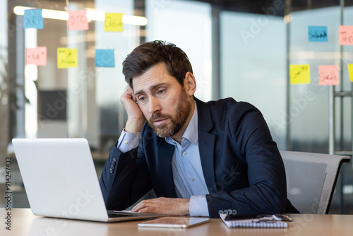 Businessman experiencing stress and boredom at work sitting at desk with laptop. Concept of office fatigue and work frustration.