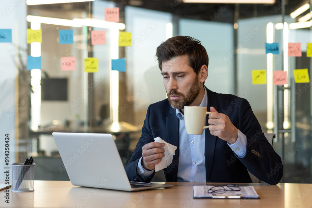 Sick businessman with tissue looking at laptop while holding coffee cup in modern office. Concept of illness at work, productivity challenges, remote work. Sick businessman with tissue looking at laptop while holding coffee cup in modern office. Concept of illness at work, productivity challenges, remote work.