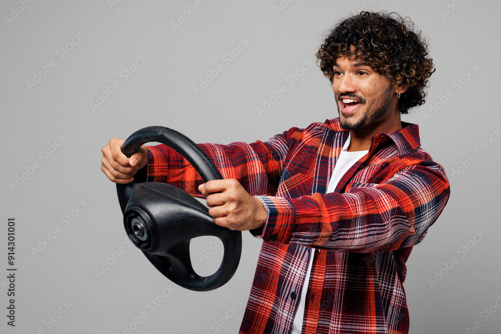 Side view young happy smiling cheerful Indian man wears red shirt casual clothes hold steering wheel driving car look aside isolated on plain grey color background studio portrait. Lifestyle concept. Side view young happy smiling cheerful Indian man wears red shirt casual clothes hold steering wheel driving car look aside isolated on plain grey color background studio portrait. Lifestyle concept.