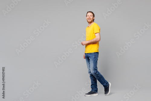 Full body side profile view smiling happy Caucasian young man he wear yellow t-shirt casual clothes looking camera walking going isolated on plain grey background studio portrait. Lifestyle concept.