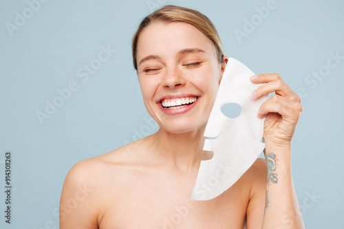 Smiling happy half naked young woman with nude make up hold apply hydrating tissue fabric textile face mask isolated on plain blue color background. Skin care healthcare cosmetic procedures concept.