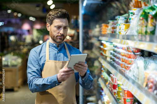 Grocery store employee with apron using tablet for inventory management in food aisle. Worker checking stock and organizing products in supermarket. Efficient retail operations with technology photo