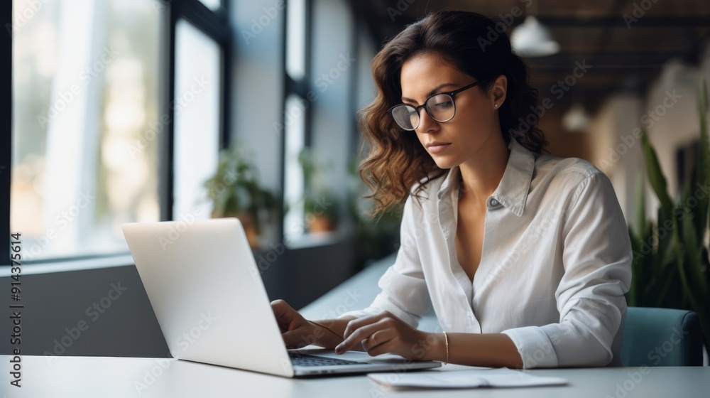 Photograph of a young woman with a determined look, working on her business plan, writing on her laptop in her minimalist studio Photograph of a young woman with a determined look, working on her business plan, writing on her laptop in her minimalist studio