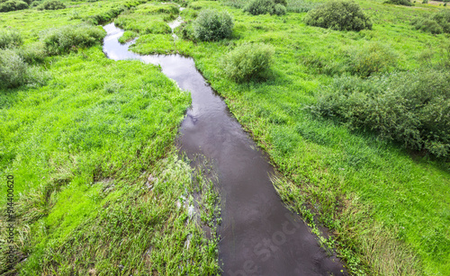 A tranquil river flows gently through vibrant green meadows, surrounded by lush vegetation under a vast blue sky.