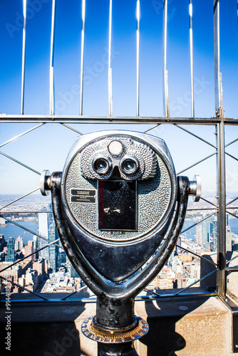 Photograph taken from the top of the Empire State Building, featuring a metallic viewing telescope with Manhattan's cityscape visible through the metal safety railings in the background