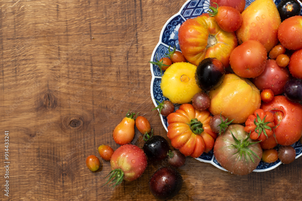 Many garden tomatoes in different colors, shapes and varieties on a blue rustic plate on a wooden table. Tomatoe composition with space for text.