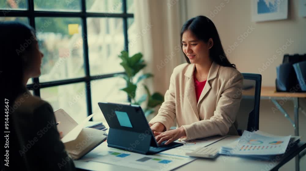 Young professional woman engages in a business meeting with a colleague in a modern office. They discuss work and review documents on a laptop and printed charts Young professional woman engages in a business meeting with a colleague in a modern office. They discuss work and review documents on a laptop and printed charts