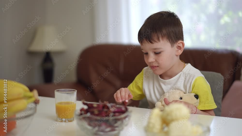 A little boy with a teddy bear in his hands pushes a glass of orange juice away from him, sitting on a chair at the table in the living room. The child ponders and smilingly takes a candy from the A little boy with a teddy bear in his hands pushes a glass of orange juice away from him, sitting on a chair at the table in the living room. The child ponders and smilingly takes a candy from the