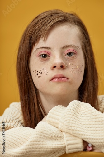 Portrait of young woman with Down syndrome having glitter on her cheeks and wearing white sweater, set against bright yellow background, resting head on hands photo