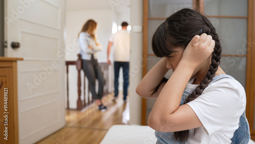 Stressed and unhappy young girl huddle in corner, cover her ears blocking sound of her parent arguing in background. Domestic violence at home and traumatic childhood develop to depression. Synchronos