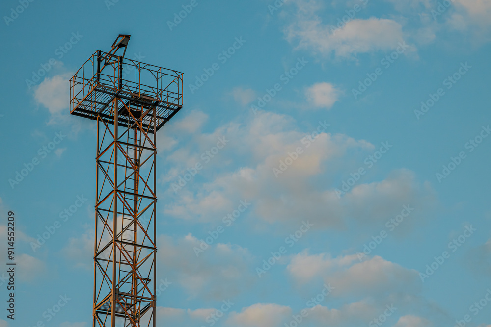 A tall metal tower with an observation platform stands against a clear blue sky dotted with soft clouds. The structure, made of metal beams, contrasts sharply with the tranquil sky A tall metal tower with an observation platform stands against a clear blue sky dotted with soft clouds. The structure, made of metal beams, contrasts sharply with the tranquil sky