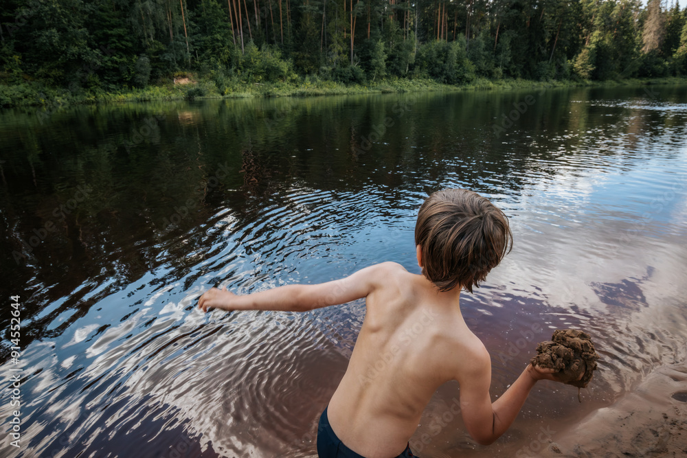 Fototapeta premium A young boy, standing at the rivers edge, is captured mid motion as he throws a rock into the calm water. The scene is set against a lush, green forest reflecting on the waters