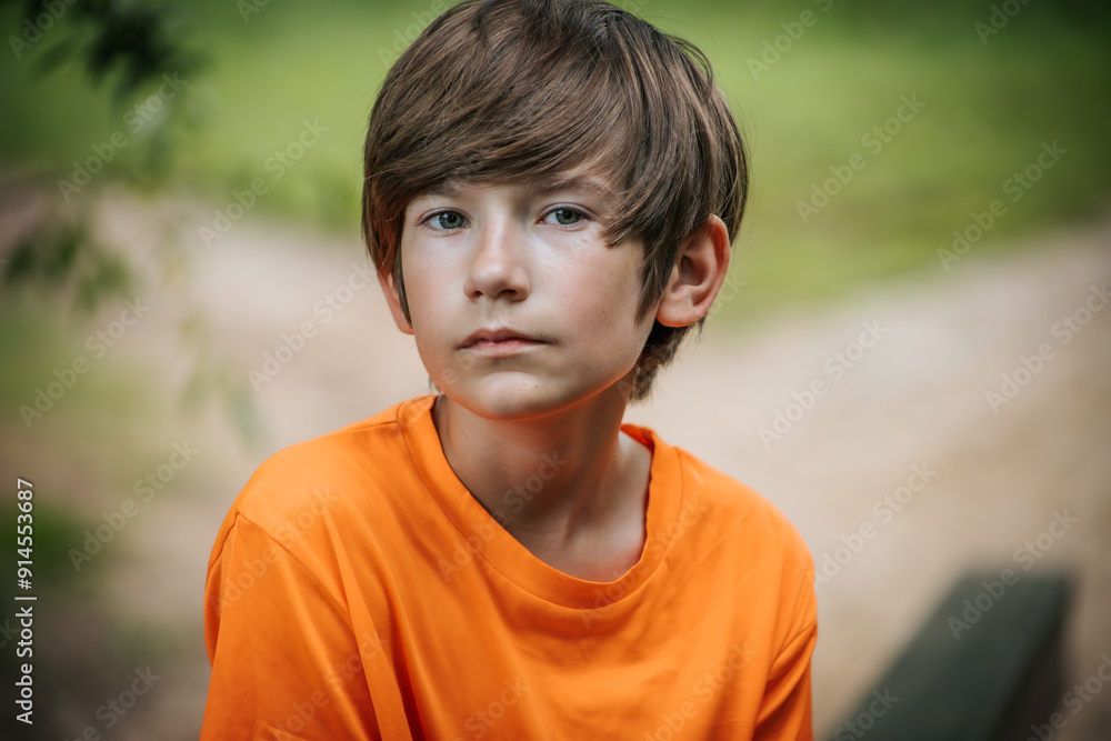 A young boy with a serious expression sits outdoors, wearing an orange shirt. The background features soft greenery, creating a serene and natural setting. A young boy with a serious expression sits outdoors, wearing an orange shirt. The background features soft greenery, creating a serene and natural setting.
