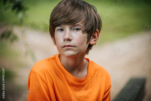 A young boy with a serious expression sits outdoors, wearing an orange shirt. The background features soft greenery, creating a serene and natural setting.