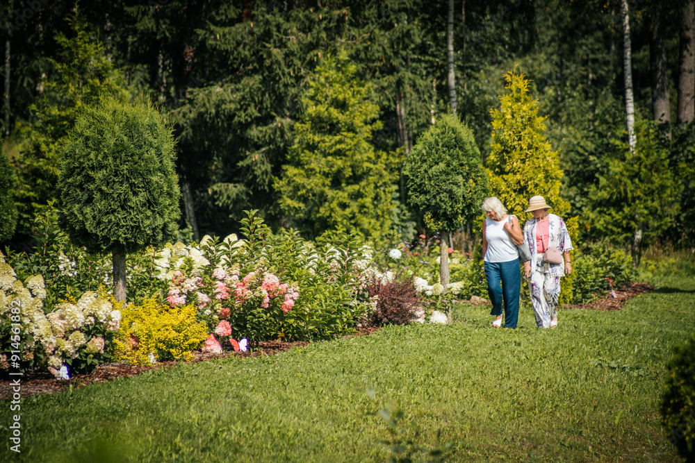 Fototapeta premium The women admire the colorful garden filled with a variety of flowers, including red, purple and yellow flowers, against a backdrop of greenery and white hydrangeas.