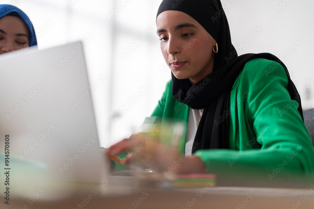 A woman in a black hijab and green blazer works on her laptop at a desk. She appears focused and engaged, highlighting a productive and professional office setting. A woman in a black hijab and green blazer works on her laptop at a desk. She appears focused and engaged, highlighting a productive and professional office setting.