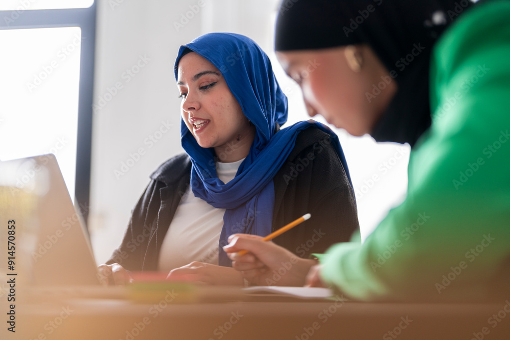 Two women in hijabs collaborate at a desk with laptops, one wearing a ...