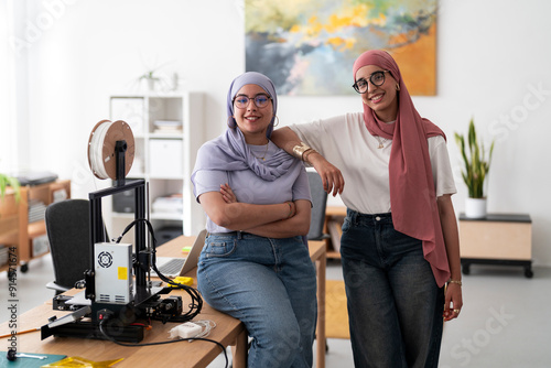 Two women in hijabs pose confidently in a 3D printing lab, surrounded by equipment and tools. Their stance reflects confidence and pride in their work and achievements in technology.