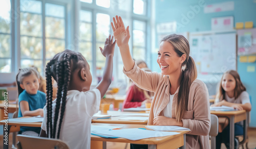 Female teacher in blouse, light cardigan giving high-five to young student with braided hair at desk, holding papers in bright classroom with natural light and students engaged in activities