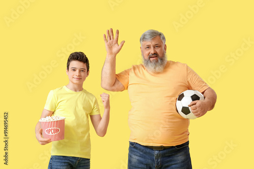 Happy grandfather with his cute grandson holding soccer ball and popcorn on yellow background