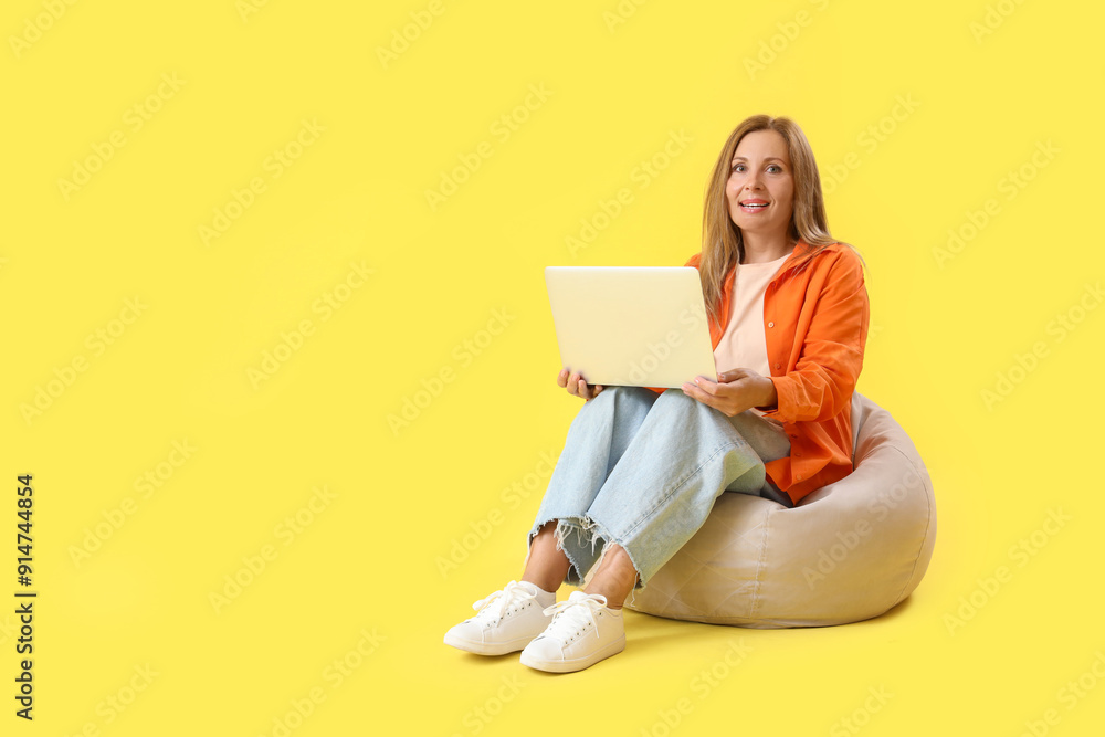 Mature woman with laptop sitting on beanbag against yellow background Mature woman with laptop sitting on beanbag against yellow background