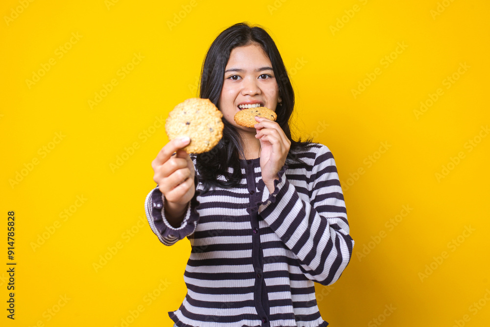 Friendly Young Asian Woman Eating While Showing Cookies Isolated On Yellow Background Friendly Young Asian Woman Eating While Showing Cookies Isolated On Yellow Background