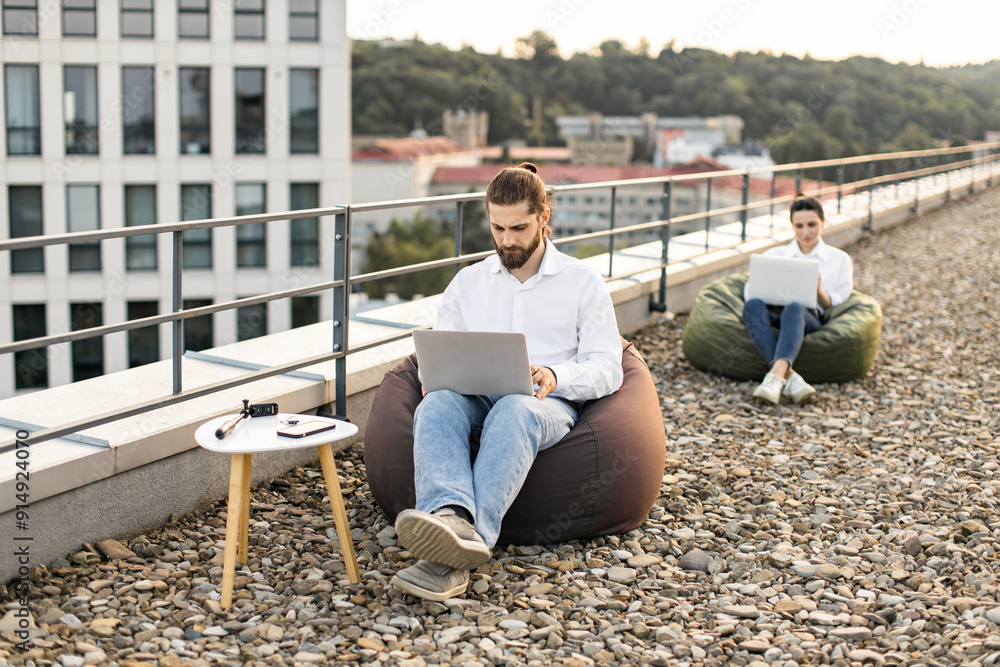 Two people working on laptops on rooftop with bean bags in urban environment. Casual outdoor workspace with urban view. Remote work, technology, relaxation, and productivity concept. Two people working on laptops on rooftop with bean bags in urban environment. Casual outdoor workspace with urban view. Remote work, technology, relaxation, and productivity concept.