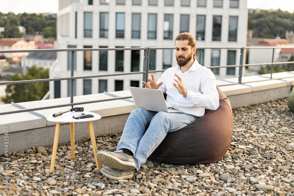 Man in casual attire working remotely on laptop outdoors. Person is seated on bean bag on rooftop terrace using laptop for communication. Scenic cityscape in background. Work-from-home concept. Man in casual attire working remotely on laptop outdoors. Person is seated on bean bag on rooftop terrace using laptop for communication. Scenic cityscape in background. Work-from-home concept.