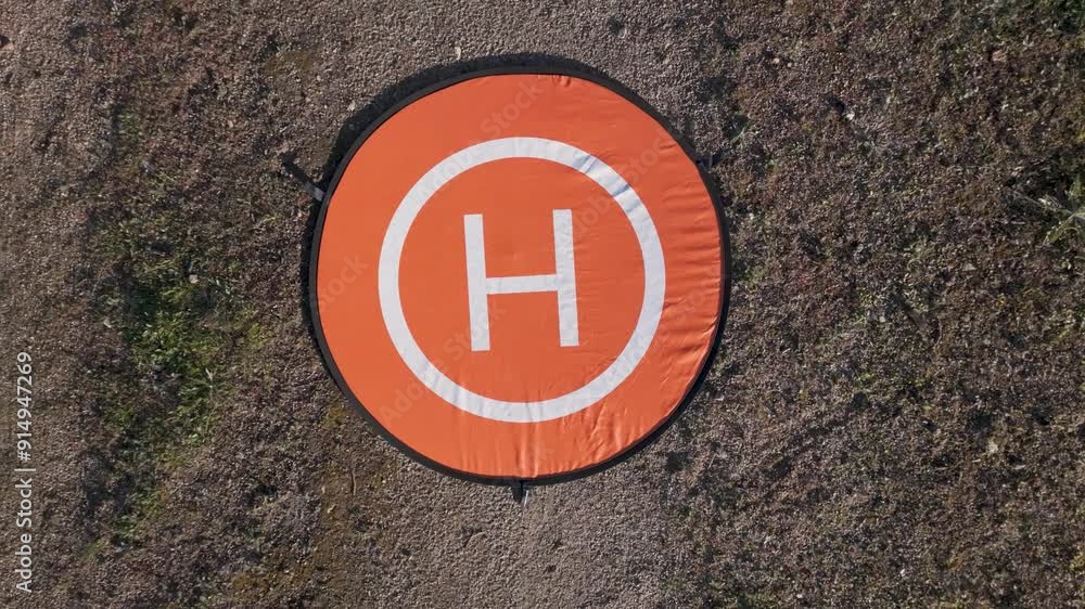 filming with a drone with a top-down view in ascending flight where we see its mobile take-off platform in orange with a black border, in white there is an inner circle and an H, the outside is land filming with a drone with a top-down view in ascending flight where we see its mobile take-off platform in orange with a black border, in white there is an inner circle and an H, the outside is land
