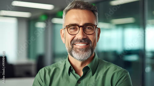 Portrait of a smiling middle-aged businessman wearing a green shirt and glasses standing in a modern office, looking at the camera with a happy expression. 