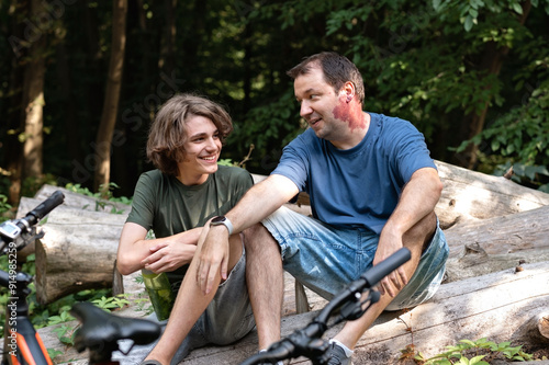 Father and teenager son sitting in forest, talking and smiling during walk in nature outdoor