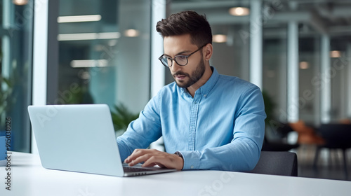 portrait of a businessman working on a laptop in a modern office, wearing a blue shirt and glasses. 